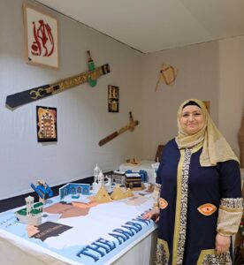 Kuvassa on pöydällä arabimaailman kartta, seinälle ripustettuna pari miekkaa. Kuvan oikeassa laidassa seisoo hymyilevä nainen.